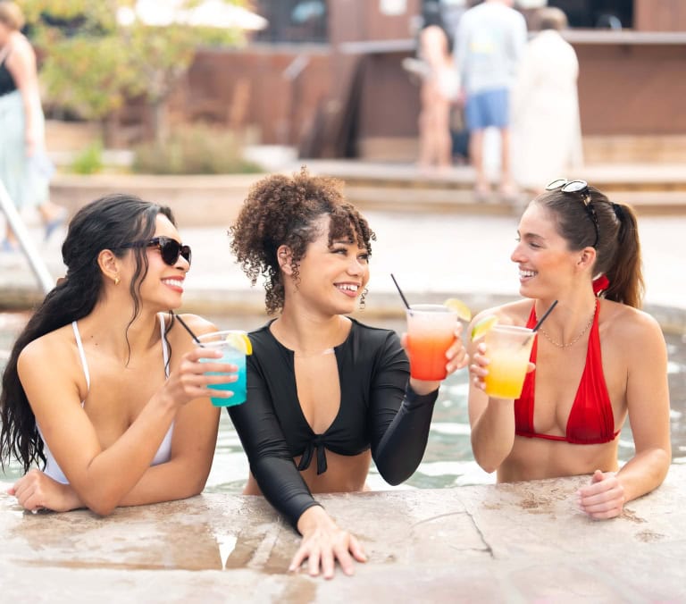 Three women, smiling, hold colorful cocktails (blue, orange, yellow) while socializing in a pool, leaning on its wet edge. Others are visible in the blurred background.