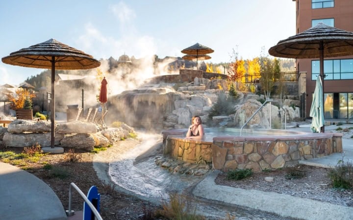 A close-up of a steaming natural hot spring pool with a thatched umbrella and autumn trees in the background.