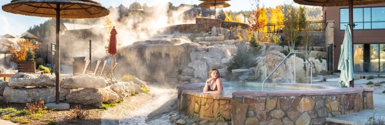A close-up of a steaming natural hot spring pool with a thatched umbrella and autumn trees in the background.