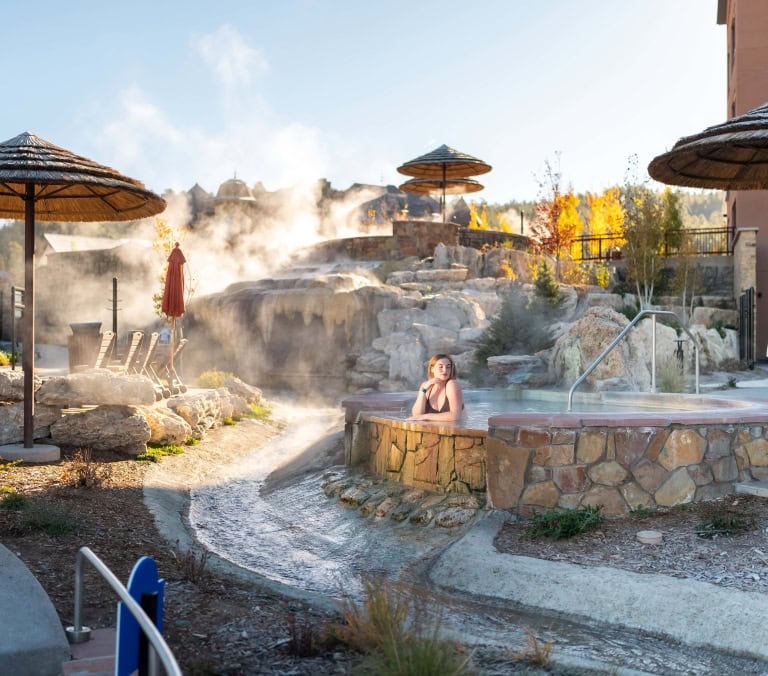 A close-up of a steaming natural hot spring pool with a thatched umbrella and autumn trees in the background.