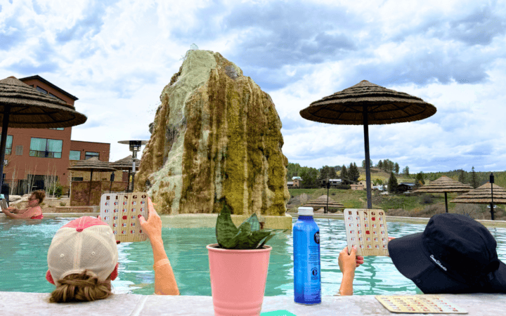 Guests relaxing in hot spring pool with rock formation and umbrellas nearby.