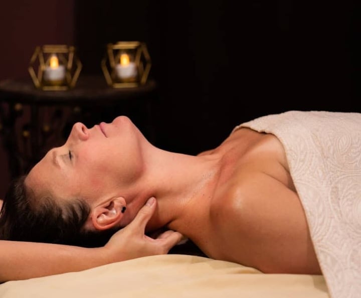 A woman receives a neck massage, eyes closed, lying draped in a towel on a table. Two glowing candles in geometric holders illuminate the dark background.
