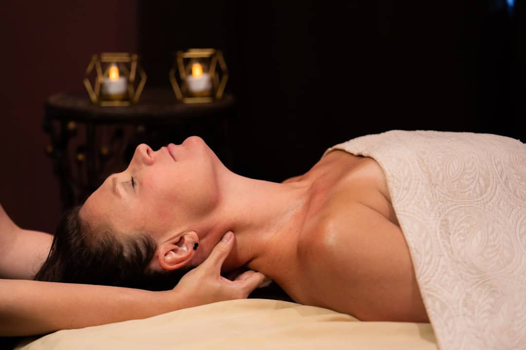 A woman receives a neck massage, eyes closed, lying draped in a towel on a table. Two glowing candles in geometric holders illuminate the dark background.