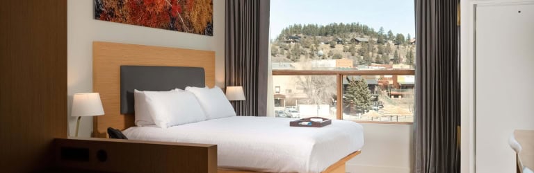 Modern hotel room with a king bed, colorful abstract art, and a large window overlooking a scenic mountain hillside.